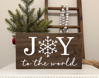 May include: A wooden sign with the words "JOY" and "to the world" in white script, with a snowflake replacing the "O". The sign is dark brown and has a small evergreen arrangement and a decorative ladder in the background.