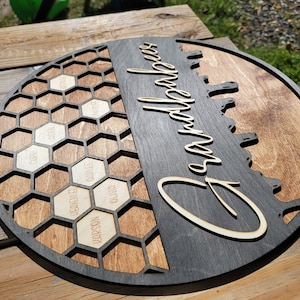 Pu&ograve; includere: Un cartello rotondo in legno con un design a nido d'ape sul lato sinistro e la parola "Grandparents" scritta in corsivo sul lato destro. Il design a nido d'ape ha celle individuali con nomi scritti al loro interno.