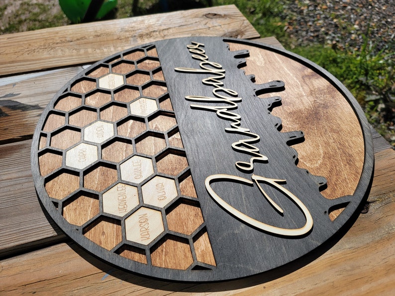 Pu&ograve; includere: Un cartello rotondo in legno con un design a nido d'ape sul lato sinistro e la parola "Grandparents" scritta in corsivo sul lato destro. Il design a nido d'ape ha celle individuali con nomi scritti al loro interno.