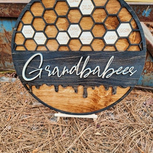 Pu&ograve; includere: Un cartello rotondo in legno con un design a nido d'ape nero e la parola "Grandbabes" scritta in corsivo bianco. Le celle del nido d'ape sono riempite con piccoli cerchi di legno con nomi scritti su di essi, tra cui "Cade", "Camilo", "Ellisyn", "Emma Kate", "Grayson", "Lucas", "Logan", "Luna" e "Sylas".