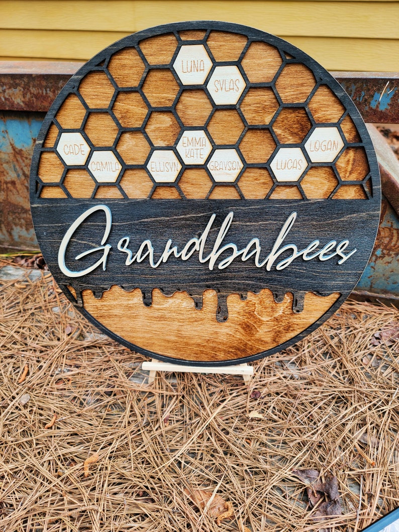 Pu&ograve; includere: Un cartello rotondo in legno con un design a nido d'ape nero e la parola "Grandbabes" scritta in corsivo bianco. Le celle del nido d'ape sono riempite con piccoli cerchi di legno con nomi scritti su di essi, tra cui "Cade", "Camilo", "Ellisyn", "Emma Kate", "Grayson", "Lucas", "Logan", "Luna" e "Sylas".