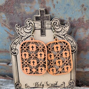 May include: A pair of wooden earrings with a cross pattern. The earrings are orange and feature a cross design with a filigree pattern. The earrings are displayed on a wooden stand with the words "Holy Script" written on it.