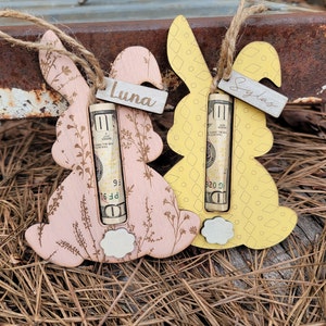 May include: Two wooden bunny ornaments with a dollar bill inserted into each. The bunny on the left is pink with a floral design and the name "Luna" written on it. The bunny on the right is yellow with a diamond pattern and the name "Sylas" written on it.