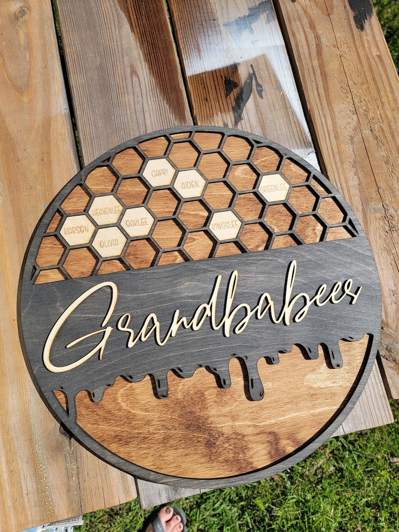 Pu&ograve; includere: Un cartello rotondo in legno con sfondo nero e un design a nido d'ape. Il cartello dice "Grandbabbees" in un font di miele che cola. Il design a nido d'ape ha nomi in ogni cella: Carson, Olivia, Heavenlee, Oaklee, Capri, Biden, Greenslee, Kingslee.