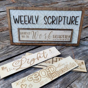 Puede incluir: Un letrero de madera con el texto "Weekly Scripture" y un letrero más pequeño con el texto "Whatever you do, work heartily - Col 3:23". Hay cinco letreros de madera más pequeños con varios versículos bíblicos, que incluyen "Be the light", "With God nothing is impossible" y "Light it"