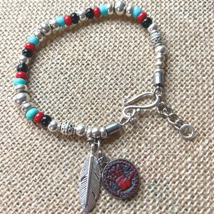 May include: A beaded bracelet featuring alternating turquoise, red, and black beads interspersed with silver-toned metal beads. The bracelet includes a silver-toned feather charm and a circular charm with a red handprint design and a hook clasp.