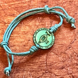 May include: A teal green bracelet with a round wooden charm. The charm has the text "SEAL OF THE KICKAPOO NATION OKLAHOMA" around the edge and a green image of a turtle with a feather in its mouth in the center.