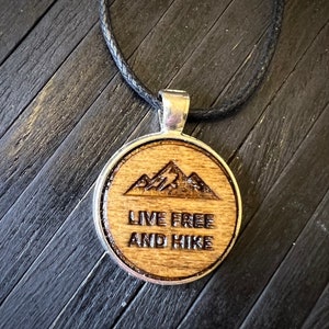 May include: A wooden pendant necklace with a black cord. The pendant is round and features a wood-burned design of mountains and the text "Live Free and Hike".