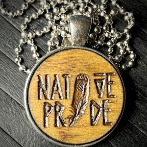 May include: A wooden pendant necklace with a silver chain. The pendant is oval shaped and has the words "Native Pride" engraved on it. A stylized feather is also engraved on the pendant.