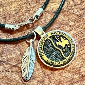 May include: A silver feather charm hangs from a black leather cord necklace with a silver clasp. The main pendant is a round, silver and gold medallion with the words "Comanche Nation Lords of the Southern Plains" engraved around the edge. The center of the medallion features a stylized image of a Native American on horseback.
