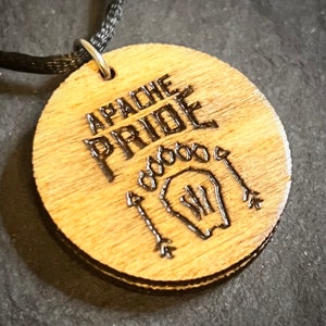 May include: A wooden pendant necklace with a black cord. The pendant is round and has the words "Apache Pride" engraved on it. The pendant also features a stylized image of a handprint and arrows.