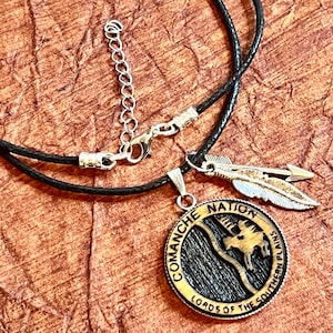May include: A black leather cord necklace with a silver pendant featuring a circular design with the text "Comanche Nation Lords of the Southern Plains". The pendant has a black and white image of a mountain range. The necklace also includes a silver feather charm.