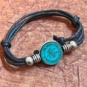 May include: Black braided cord bracelet with a silver metal bead and a round teal blue charm with the words "The Great Seal of the Choctaw Nation" in silver lettering.