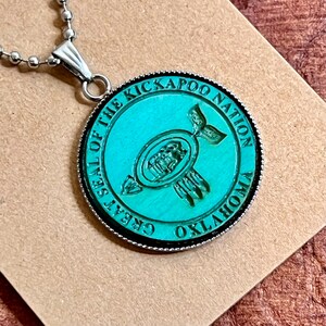 May include: A teal blue pendant necklace with a silver chain. The pendant features the seal of the Kickapoo Nation of Oklahoma. The seal depicts a stylized Native American design with a circle and feathers.