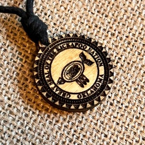 May include: A wooden pendant necklace with a black cord. The pendant is circular with a detailed, engraved design. The text "GREAT SEAL OF THE KICKAPOO NATION OKLAHOMA" surrounds a central emblem. The pendant is light brown with dark brown engraved details.
