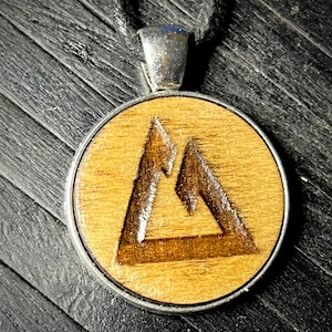 May include: A wooden pendant with a silver-toned metal frame and a carved mountain design. The pendant is on a black cord.