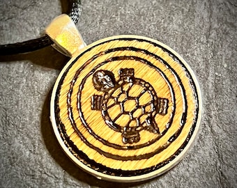 May include: A wooden pendant necklace with a black cord. The pendant is round and features a wood-burned design of a turtle.