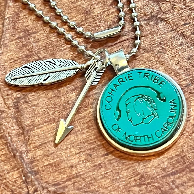 May include: A silver chain necklace with a green circular pendant featuring the text "Coharie Tribe of North Carolina". The pendant has a stylized image of a Native American person's head. The necklace also has a silver arrow and feather charm.