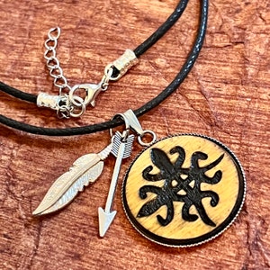 May include: A black leather cord necklace with a wooden pendant featuring a black and white design. The pendant is surrounded by a silver-colored metal rim. The necklace also includes a silver-colored feather charm and arrow charm.