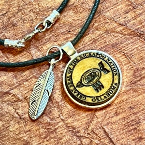 May include: A black leather cord necklace with a silver feather charm and a round wooden pendant. The pendant features the Great Seal of the Kickapoo Nation, Oklahoma, with a stylized image of a bird and the words "Great Seal of the Kickapoo Nation Oklahoma".