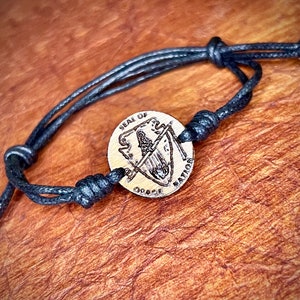May include: A black cord bracelet with a wooden charm featuring the text "SEAL OF OATH" and "NATION".