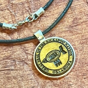 May include: A wooden pendant necklace with a black cord. The pendant features the seal of the Kickapoo Nation, Oklahoma. The seal depicts a stylized bird with a handprint design.