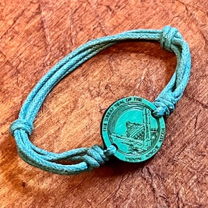 May include: A teal blue braided cord bracelet with a round metal charm. The charm has the words "The Great Seal of the" and "State of Utah" engraved on it.