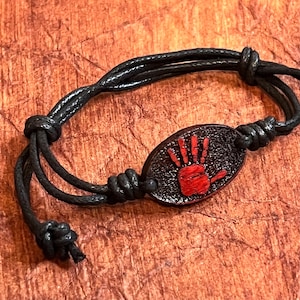 May include: Black cord bracelet with a red handprint charm. The handprint is on a black oval-shaped charm.