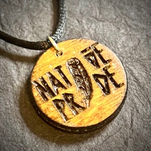 May include: A wooden pendant necklace with a black cord. The pendant is round and has the words "NATIVE PRIDE" carved into it.