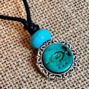 May include: A turquoise and silver necklace with a pendant featuring a stylized fish design. The pendant is surrounded by a decorative silver frame.