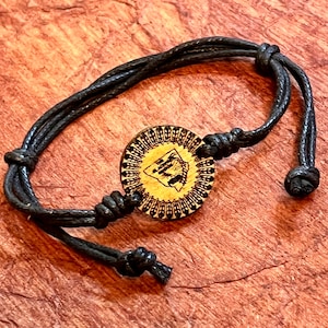 May include: A black cord bracelet with a wooden charm featuring a detailed outline of a state. The charm has a decorative border.