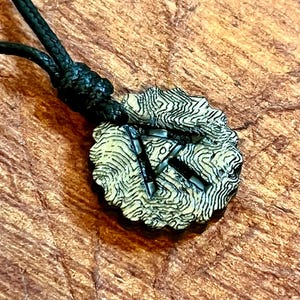May include: A black cord necklace with a round pendant featuring a stylized design in black and white. The pendant has a textured surface with a swirling pattern.