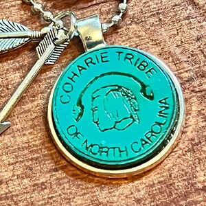 May include: A green and silver necklace with a round pendant featuring the text "Coharie Tribe of North Carolina" and a stylized image of a Native American person's head. The pendant is attached to a silver chain with two silver arrow charms.