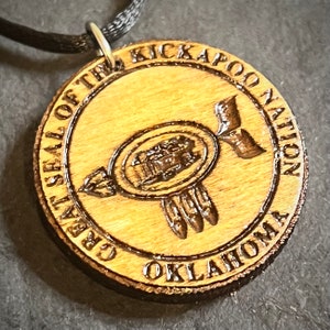 May include: A wooden pendant with a brown cord. The pendant is a round disc with the words "Great Seal of the Kickapoo Nation Oklahoma" engraved around the edge. In the center of the disc is a stylized image of a bird with feathers.