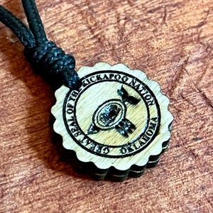 May include: Wooden pendant necklace featuring the seal of the Kickapoo Nation, Oklahoma. The round pendant has a scalloped edge and is attached to a black cord. The seal is engraved in black.