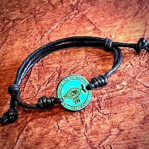 May include: Black cord bracelet with a turquoise blue metal charm featuring the text "SEAL OF THE KICKAPOO TRIBE OKLAHOMA".