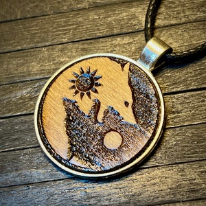 May include: A wooden pendant necklace with a sun and moon design. The pendant is round and has a silver-colored metal rim. The sun is on the light wood side and the moon is on the dark wood side. The pendant is hanging from a black cord.