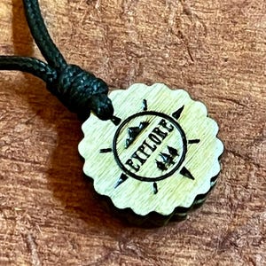 May include: Wooden necklace with a round pendant featuring the word "Explore" in a compass design with mountains and trees. The pendant is attached to a black cord.