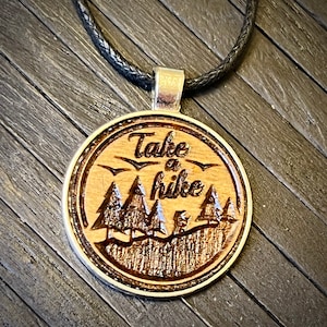 May include: A wooden pendant necklace with a black cord. The pendant is round and features a wood-burned design of trees and the words "Take a hike".