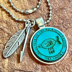 May include: A silver chain necklace with a round pendant featuring the "Great Seal of the Kickapoo Nation, Oklahoma" in turquoise blue. The pendant is attached to a silver arrow and feather charm.