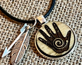 Peut inclure: Un collier pendentif avec un pendentif rond en bois représentant une main et une spirale. Un charme en argent en forme de flèche et de plume est suspendu à côté du pendentif. Le collier a un cordon noir.
