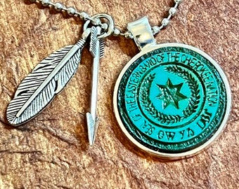 May include: A silver necklace with a green pendant featuring the text "Seal of the Eastern Band of the Cherokee Nation". The pendant also has a star and a wreath design. The necklace also has a silver arrow and feather charm.