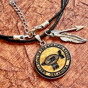 May include: A pendant necklace featuring a round wooden pendant with the words "GREAT SEAL OF THE KICKAPOO NATION OKLAHOMA" around the edge. The pendant has a black and brown emblem in the center. The necklace also includes a silver arrow and feather charm.