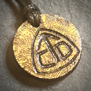 May include: A wooden pendant necklace with a black cord. The pendant is round and has a carved design of a shield with the letters "BD" inside.