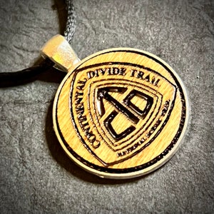 May include: A wooden pendant with the text "CONTINENTAL DIVIDE TRAIL" and a shield-shaped emblem. The pendant is round, with a light brown wood grain and a black cord. The emblem features the letters "CD" inside a shield.