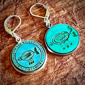 May include: A pair of silver earrings with turquoise blue circular pendants. Each pendant features a silver design of a Native American symbol with the text "Great Seal of the Kickapoo Nation Oklahoma" around the edge.