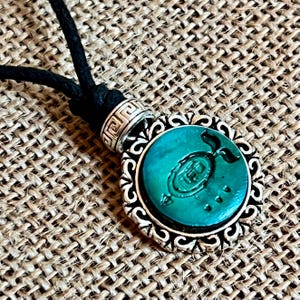 May include: A silver pendant necklace with a green gemstone cabochon featuring a stylized design. The pendant is surrounded by a decorative silver frame and hangs from a black cord.