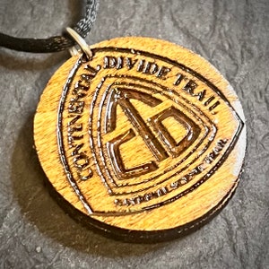May include: Wooden pendant necklace with a brown cord. The pendant is round and features a carved design of the Continental Divide Trail logo, which includes the words "Continental Divide Trail" and a stylized arrow pointing upwards.