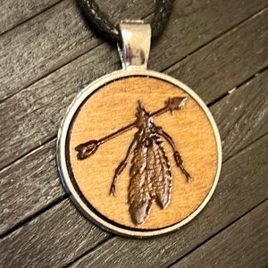 May include: A wooden pendant necklace with a silver-toned metal frame. The pendant features a wood-burned image of a single feather with an arrow through it.