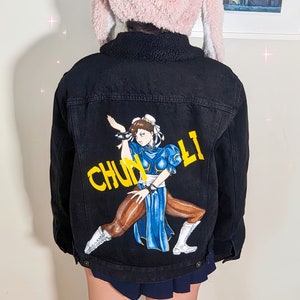May include: Black denim jacket with a hand-painted image of Chun-Li from Street Fighter. The image is in yellow, blue, and brown colors. The text "Chun-Li" is written in yellow letters.